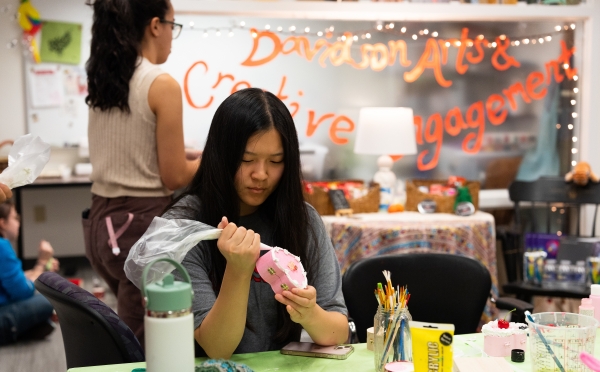 A student focused on her work uses a piping bag to apply white decorative frosting to a small heart-shaped pink jewelry box in a creative studio labeled "Davidson Arts & Creative Engagement."