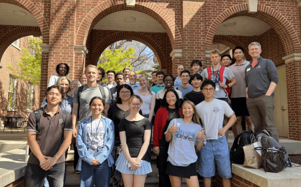 A large, diverse group of students and faculty pose together for a commemorative photo on the steps of a brick building with prominent arched entryways.