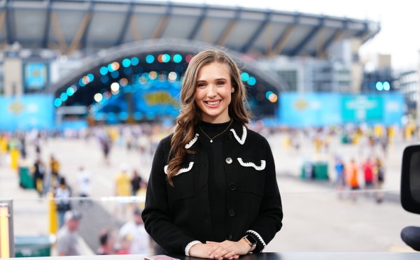 Courtney smiles at a desk with a stadium and large outdoor event stage blurred in the background.
