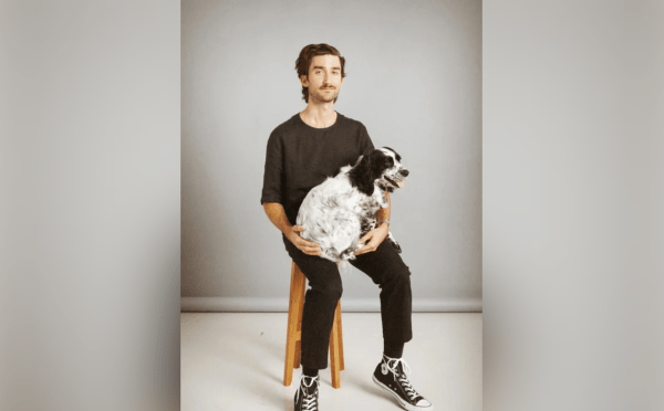 A man with a mustache sits on a wooden stool against a gray backdrop, wearing a black t-shirt, black pants, and black high-top sneakers while cradling a fluffy, black-and-white speckled dog on his lap.