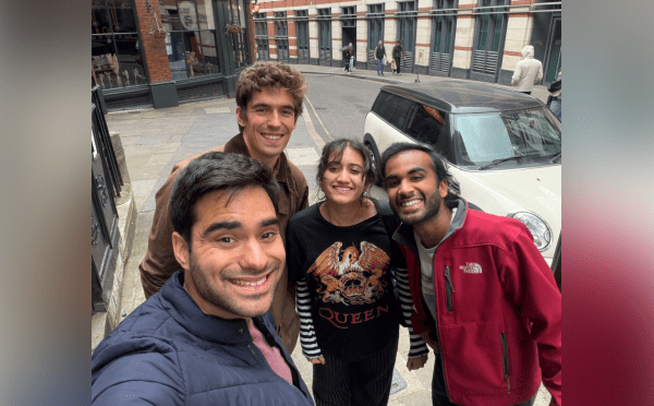 Four smiling friends take a high-angle selfie on a curved London street in front of a white compact car and a building labeled "Pub & Eating House."