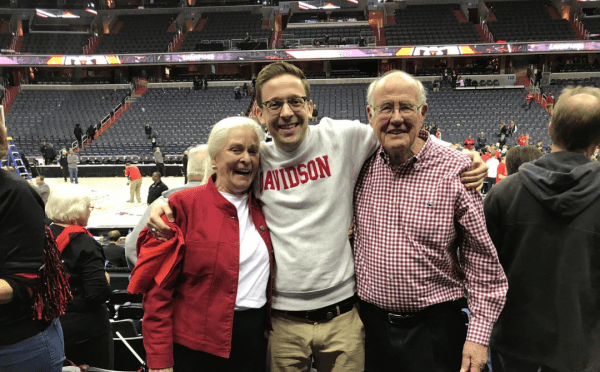 A smiling young man in a white Davidson sweatshirt stands with his arms around an older woman in a red jacket and an older man in a checkered shirt in a basketball arena.