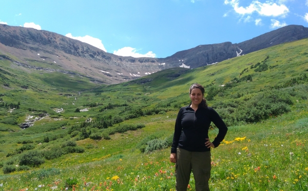 A smiling woman stands in a lush, green mountain valley filled with yellow and red wildflowers under a blue sky with light clouds.