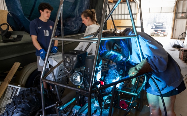 A student wearing a blue welding helmet and protective gloves welds a component onto the metal chassis of a custom off-road vehicle while two peers observe.