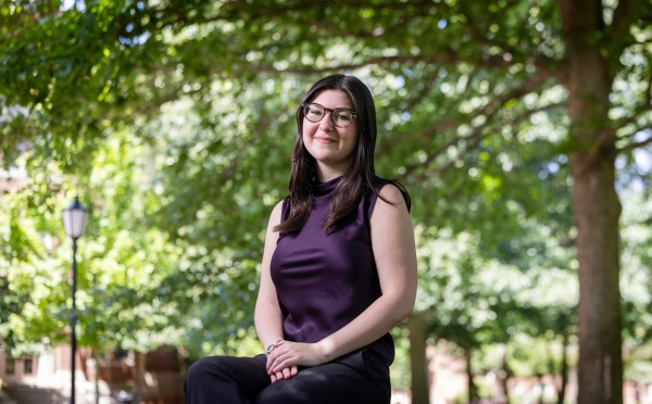 A smiling student wearing glasses and a dark purple sleeveless top sits outside on a stone ledge beneath leafy green trees.