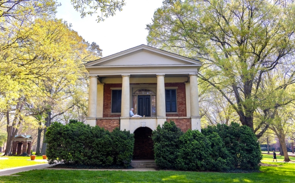 A person sits reading on the upper portico of a historic brick building with classical white columns, surrounded by leafy green trees and hedges on a sunny campus.