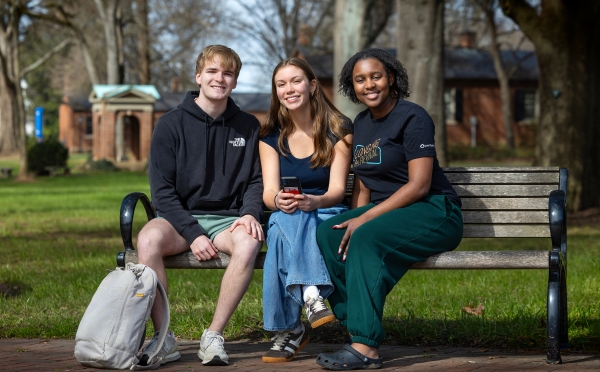 Three students sitting together on a wooden park bench, smiling at the camera.