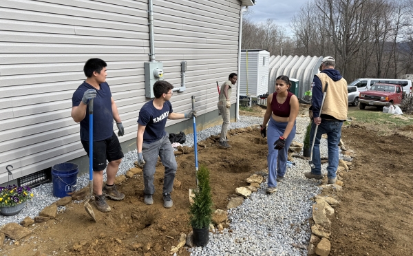 A group of volunteers works together on an outdoor landscaping project next to a house, digging trenches and laying gravel and stone borders.
