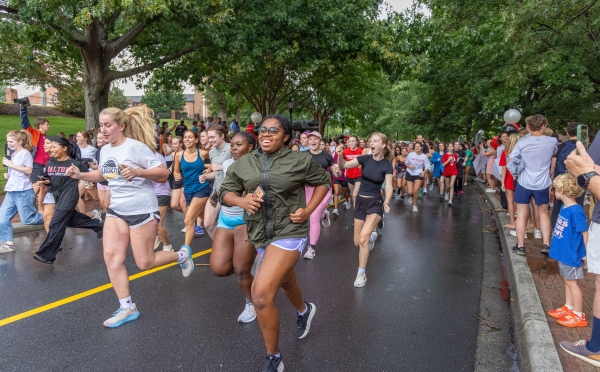 A large group of smiling young runners race down a tree-lined paved road while spectators cheer along the sidewalk.