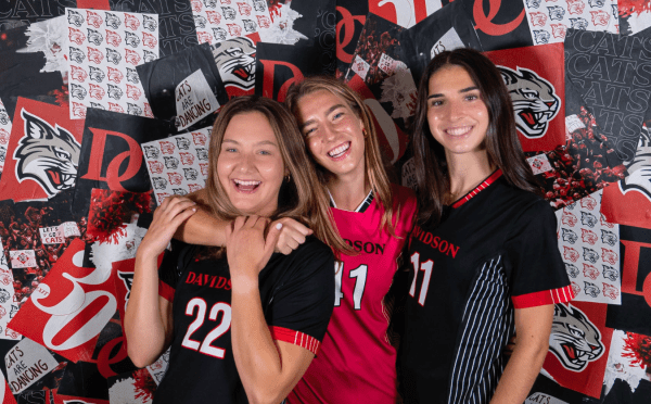 Three smiling female soccer players in Davidson College jerseys pose together in front of a red and black branded backdrop.