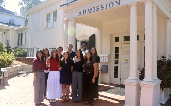 A diverse group of nine students smiling together for a group photo under the columned portico of a white "Admission" building.