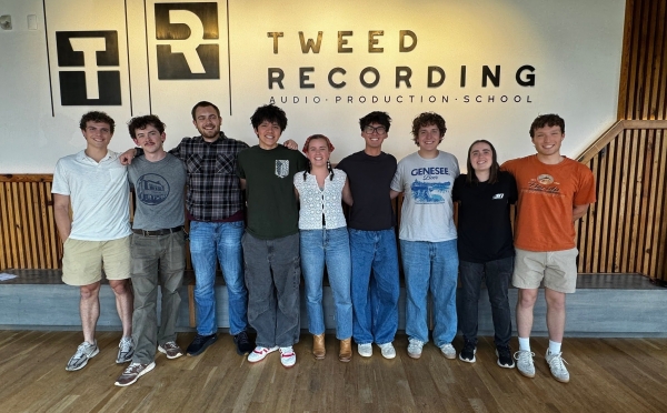A group of eight young adults stands in a line with their arms around each other's shoulders, posing for a photo inside a modern room in front of a wall with a logo that reads "Tweed Recording Audio Production School."