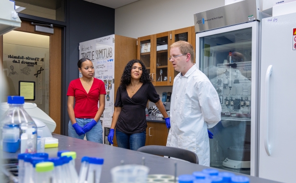 Dr. Thurtle-Schmidt, wearing a white lab coat, speaks with two student researchers wearing protective blue gloves inside a science laboratory.