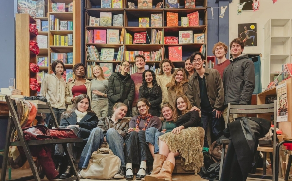 A diverse group of smiling young adults poses together inside a cozy bookstore with tall shelves filled with children's books and illustrations in the background.