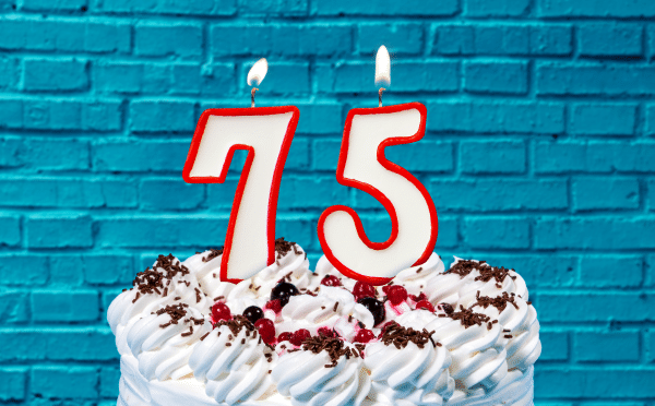 A cake in the foreground, holding candles in the shape of "75", against a blue brick wall of a retro soda shop