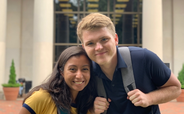 Zach and Yara smile together while wearing backpacks outdoors in front of a stone campus building with large glass windows.