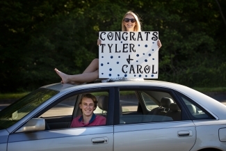 Friend sitting on top of car with sign "Congrats Tyler & Carol" 