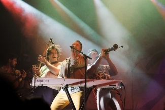 A group of three men play instruments on stage as colorful lights cast over them