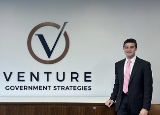 a young white man wears a suit and tie while standing in front of a wall that reads "Venture Government Strategies"