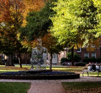 a student sits on a bench working on a laptop as a silver statue is in the background 