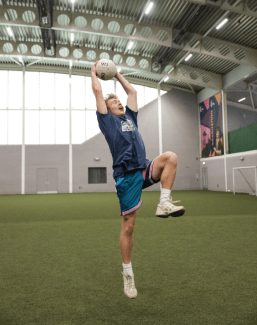 a male wearing athletics throws a soccer ball while in an indoor field