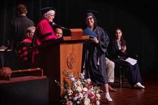 a young woman accepts an award on a stage while wearing regalia