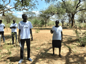 a group of young Black people stand in a grassy opening surrounded by treees