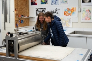 two young women stand together smiling over a printing press