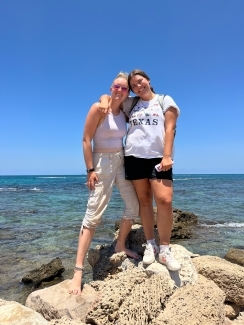 two young people stand on rocks along an ocean smiling on a sunny day