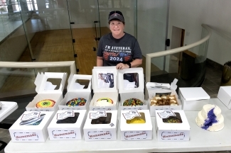 an older woman stands behind a table of cakes