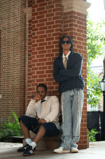 two young men stand in front of a brick wall 