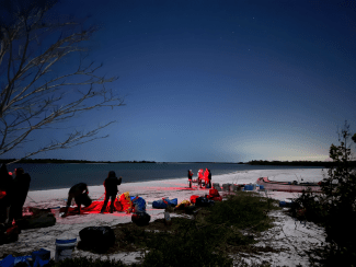 young people on a beach at sunrise using red lights to pack up camping supplies