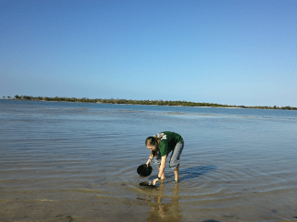 a young woman dips two pans into a body of water on a sunny day