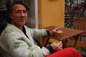 a middle aged man sits at a table outside drinking a coffee and smoking