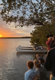 a group of students sitting on a dock looking out at a body of water at sunset