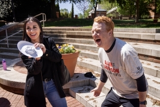 a young woman holds up a pie to a professor's face on a sunny day
