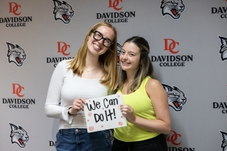 two young women smile and hold a sign that reads "we can do it"