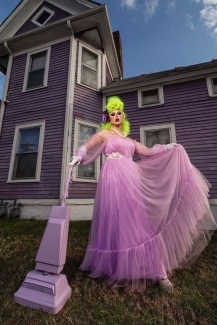 a young white woman wears drag while vacuuming in front of a purple house