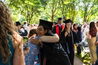 a young woman hugs a family member while wearing graduation regalia on a sunny day