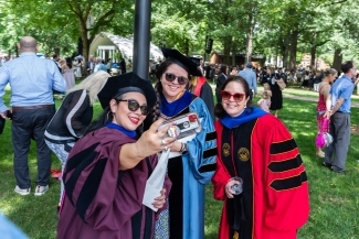 three professors pose together taking a selfie in graduation regalia on a sunny day