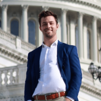 a young white man wears a jacket and stands in front of the U.S. Capitol