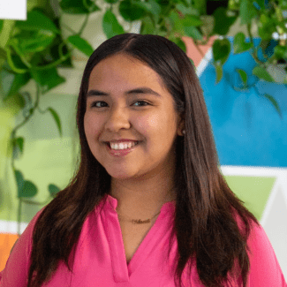 a young woman with dark hair wearing a pink stop standing in front of a colorful wall with plants in the background