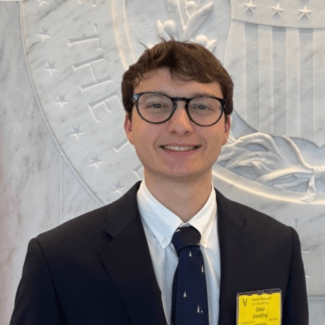 a young white man wearing glasses and a suit and tie in front of a federal seal