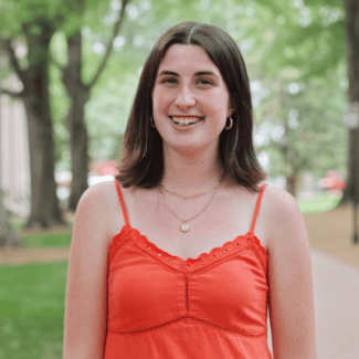 a young white woman with dark brown hair wearing an orange top smiling outside