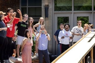 A diverse group of enthusiastic college students with expressions of excitement and surprise gather outside a brick campus building, some wearing university apparel and taking photos, as they react energetically to something happening off-camera