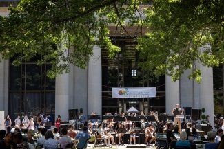 A student orchestra performs outdoors in front of a columned campus building during the Verna Miller Case Research and Creative Works Symposium, with a speaker at a podium addressing the audience while lush green trees frame the classical architecture.