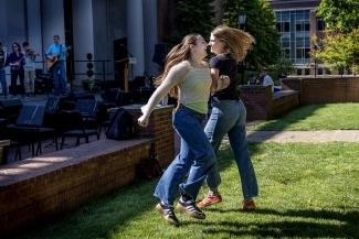 Two female students dance joyfully on a grassy lawn near a campus performance space where musicians with guitars and other instruments play on a small stage in the background