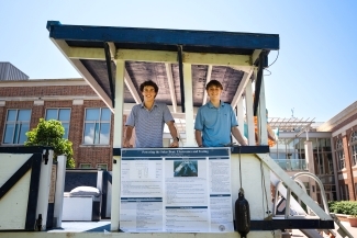 Two smiling male students in blue polo shirts stand proudly beside their solar boat project display titled "Powering the Solar Boat: Electronics and Testing," showcasing their engineering work under a small shelter with campus buildings visible in the background.