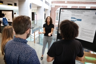 A student explains her research poster on virtual reality brain rehabilitation to a small group inside an academic building.