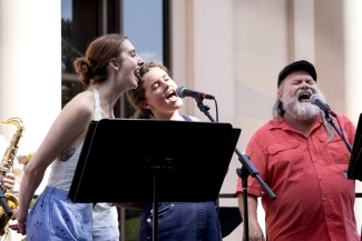 two young women sing into a microphone together while an older man sings behind them
